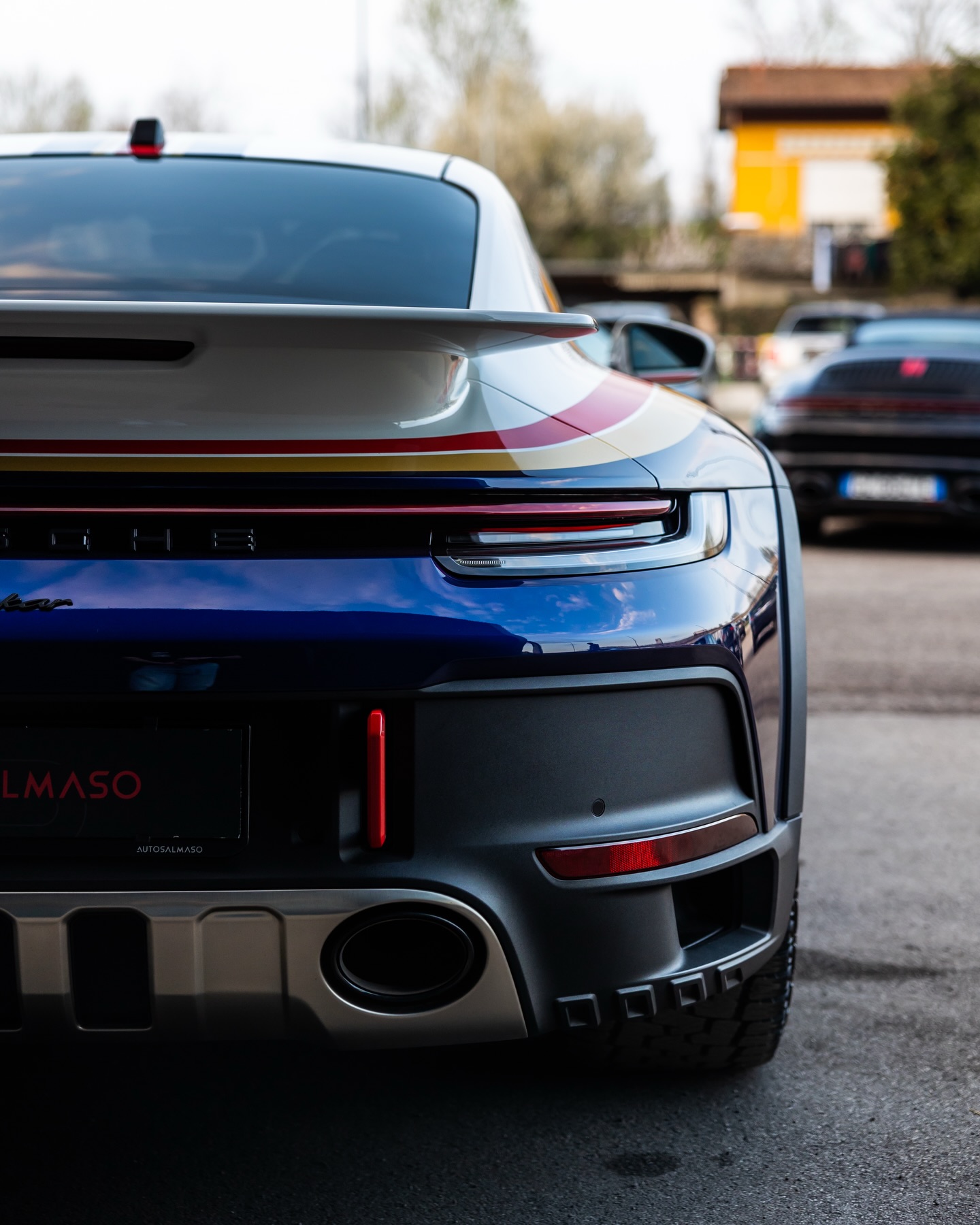 Posteriore di Porsche 911 Dakar blu e bianca con strisce rosse e oro, targa AutoSalmaso, altre auto sul piazzale