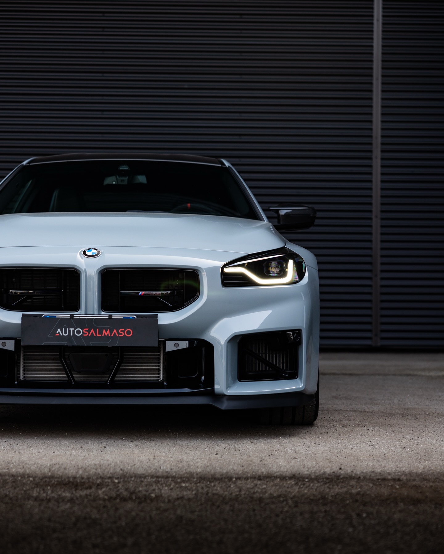 BMW M2 grigio chiaro con fari LED accesi e targa AUTOSALMASO, davanti a una saracinesca metallica scura